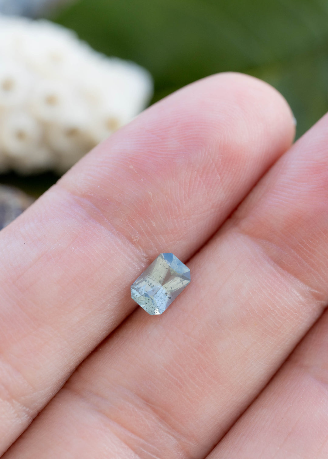 Natural smoky aqua blue Montana sapphire weighing 0.71 carats shown on fingertips to demonstrate its size, precision radiant cut, and natural inclusions.