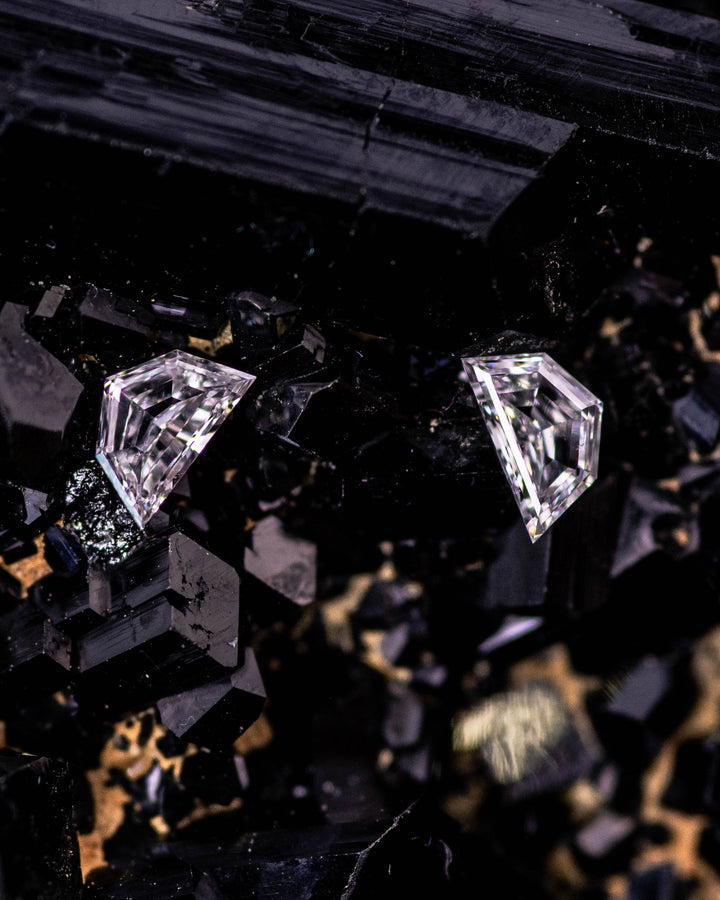 Macro photo of two 0.18 carat D color Cadillac cut diamonds from South Africa resting on dark tourmaline crystals. The natural untreated pair displays sharp geometric facets and VVS–VS clarity with mirror-like brilliance