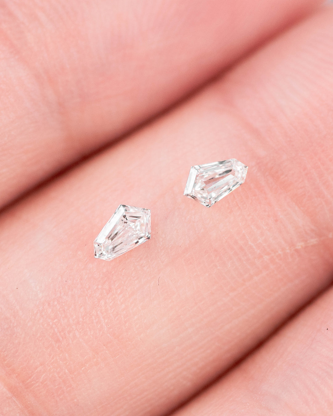 Pair of 0.19 carat F-G color VVS-VS clarity kite step cut diamonds from South Africa resting on a fingertip, showing their faceting, symmetry, and brilliance in a close-up hand view.