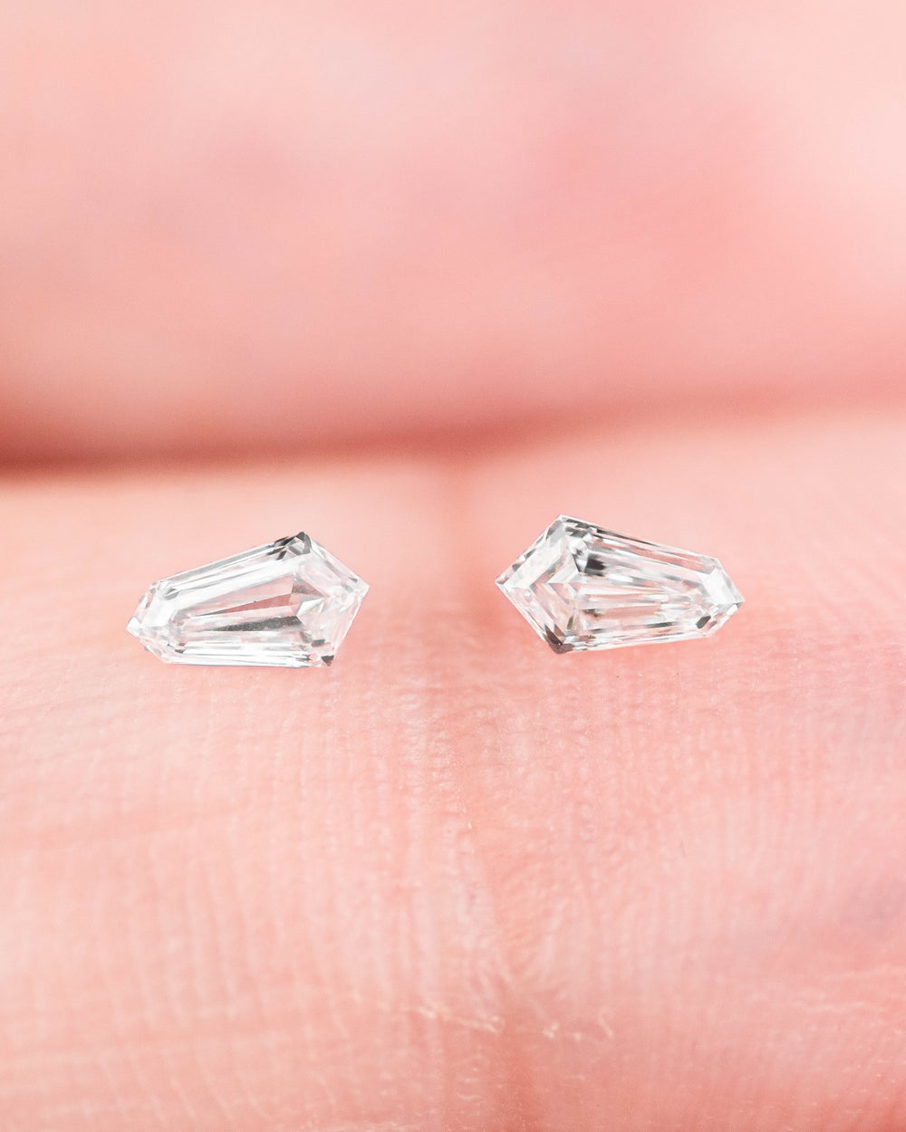 0.19-carat D-E color VVS clarity kite step-cut diamond pair from South Africa resting on fingertip, showing matched geometric shape, brilliant faceting, and high-clarity sparkle — loose diamonds for fine jewelry and custom designs.