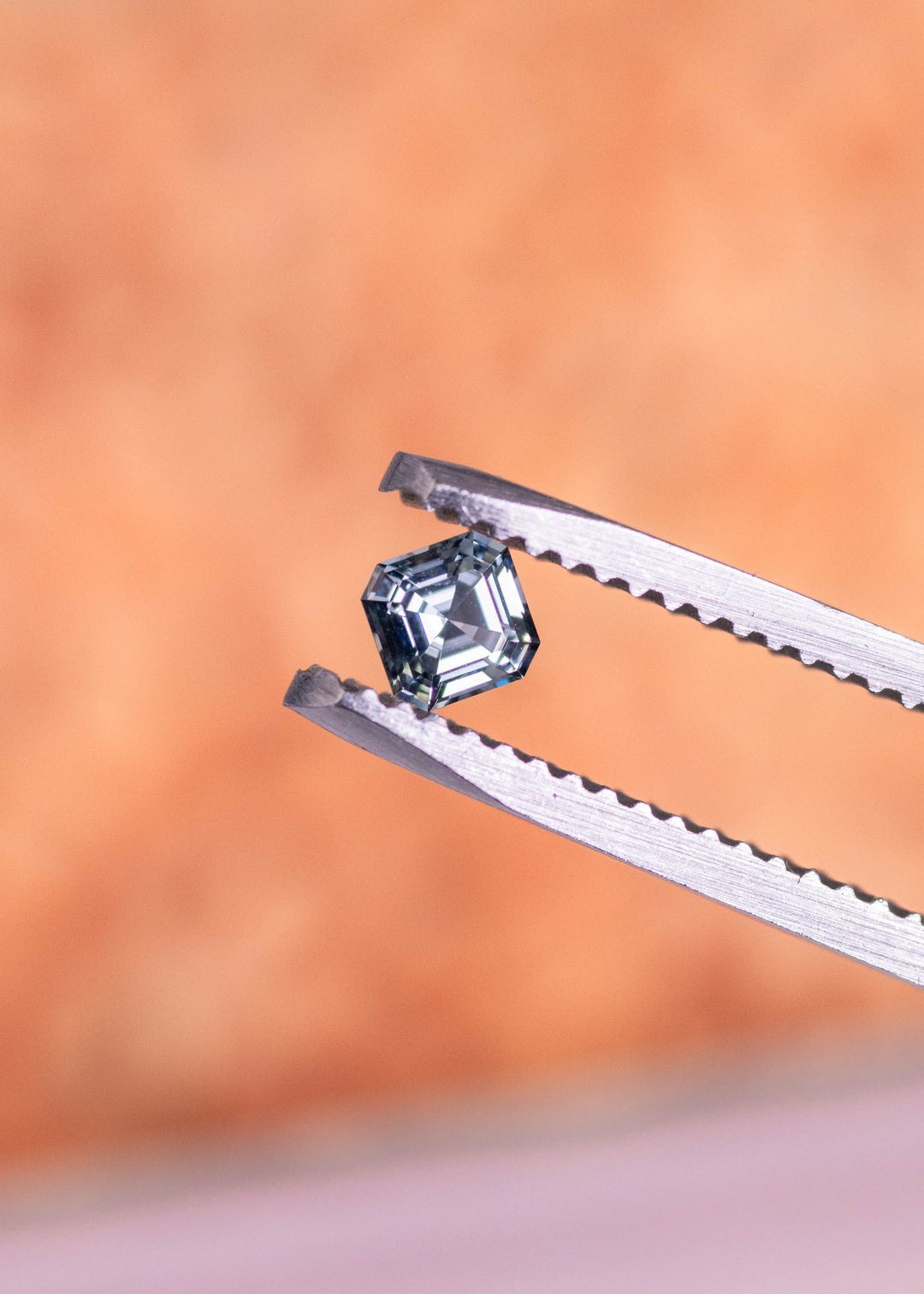 Loose blue-green Montana sapphire held in tweezers, highlighting the Asscher cut step facets and natural teal color shift as it catches the light.
