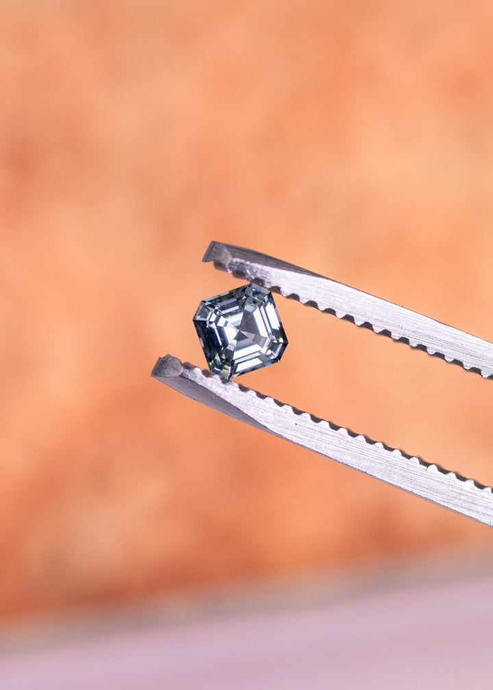 Loose blue-green Montana sapphire held in tweezers, highlighting the Asscher cut step facets and natural teal color shift as it catches the light.