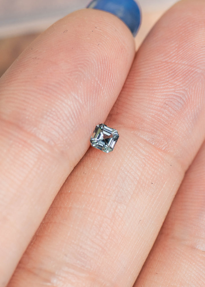Loose Montana sapphire resting on fingertips, showing the square Asscher cut and subtle shift between blue, teal, and green tones.