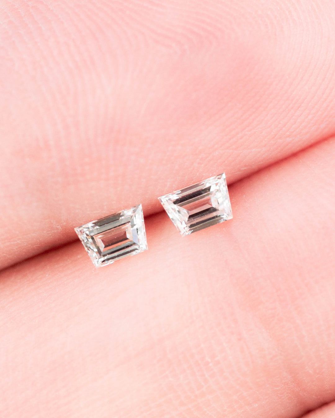 Matched pair of 0.28 carat F color VVS clarity Trapezoid step cut diamonds displayed on a fingertip, showcasing the precision faceting, brilliance, and elongated geometric shape of the loose South African diamonds.