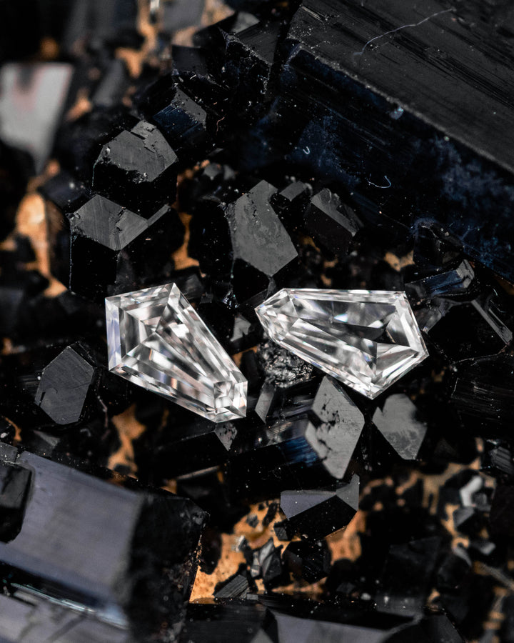 Pair of 0.28 carat G color VVS clarity kite step cut diamonds displayed on a black crystal mineral matrix, showcasing the geometric faceting, brilliance, and precision-cut shape of both stones from South Africa.
