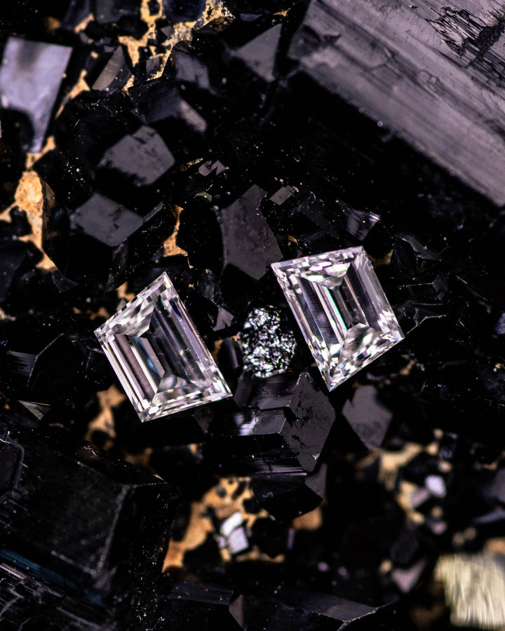 Macro photograph of 0.31 carat E–F color VVS clarity trapezoid step cut diamond pair from South Africa placed on black tourmaline crystals. The untreated diamonds showcase mirror-like precision cuts and reflective fire.