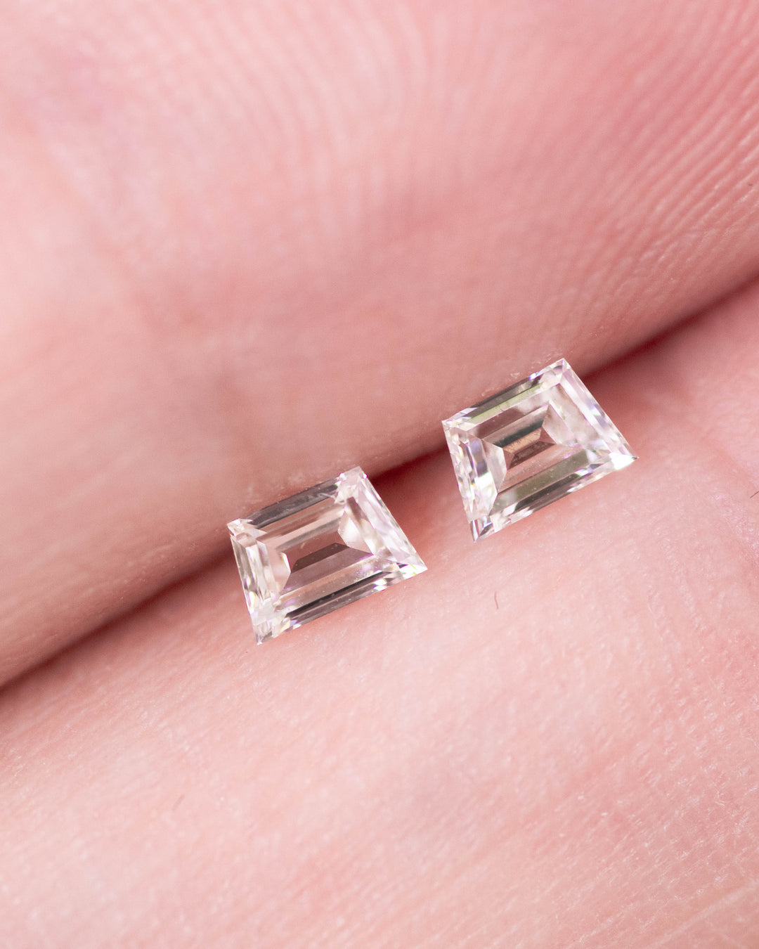 Close-up of 0.31 carat G–H color VVS clarity trapezoid step cut diamond pair from South Africa held on fingertips. The natural untreated diamonds show sharp facet lines and precise matching geometry ideal for custom fine jewelry.