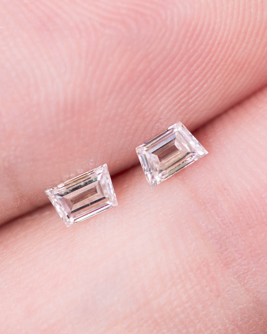 Close-up of 0.33 carat F color VVS clarity trapezoid step cut diamond pair from South Africa displayed on fingertips. The untreated diamonds showcase clean lines, flawless step faceting, and perfect matching proportions for fine jewelry settings.
