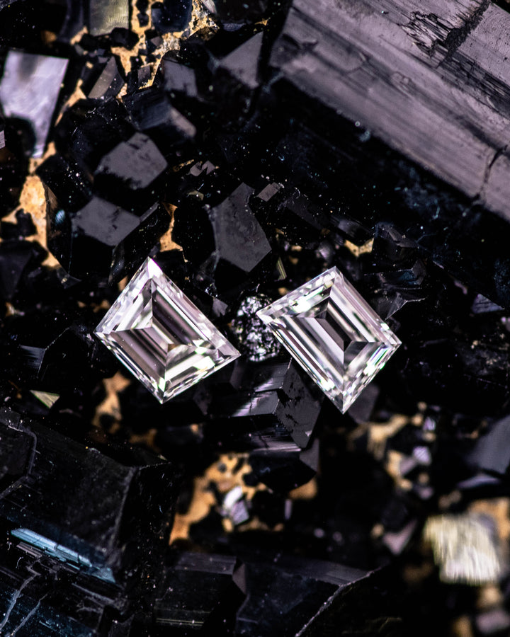 Macro image of a 0.34 carat F–G color VVS clarity trapezoid step cut diamond pair from South Africa resting on a black crystal matrix. The untreated diamonds display sharp step faceting, bright reflections, and precise symmetry.
