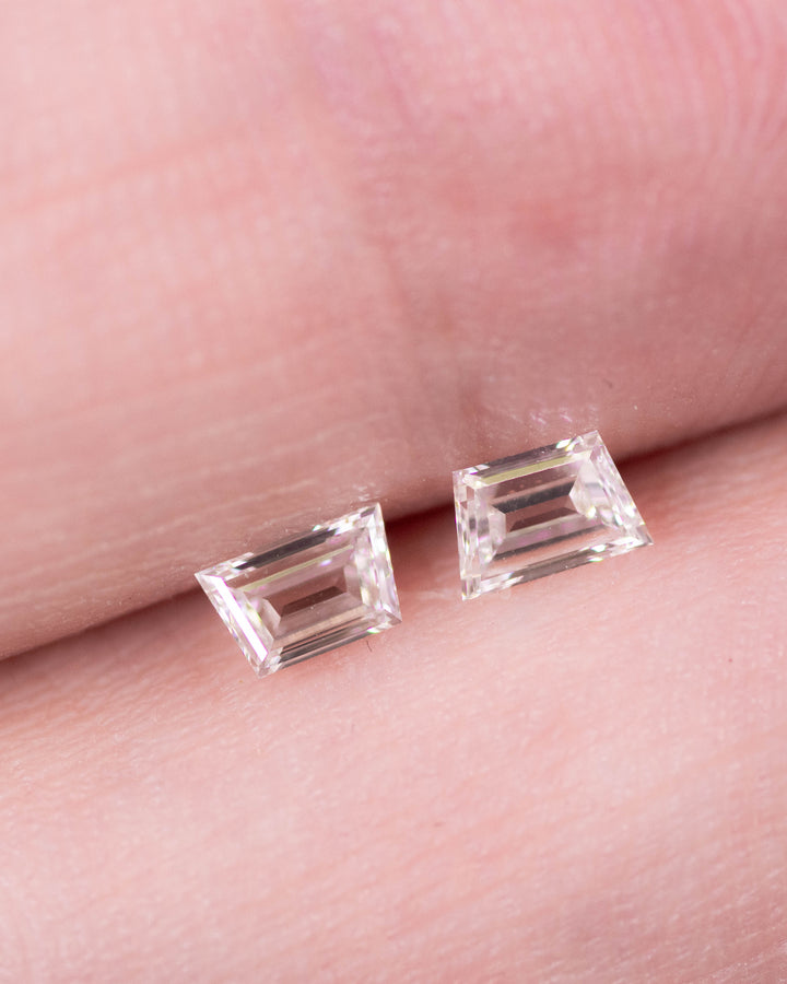 Close-up of a 0.34 carat G color VVS clarity trapezoid step cut diamond pair from South Africa displayed on fingertips. The natural untreated diamonds exhibit precise symmetry, mirror-like faceting, and fine craftsmanship ideal for jewelry settings.