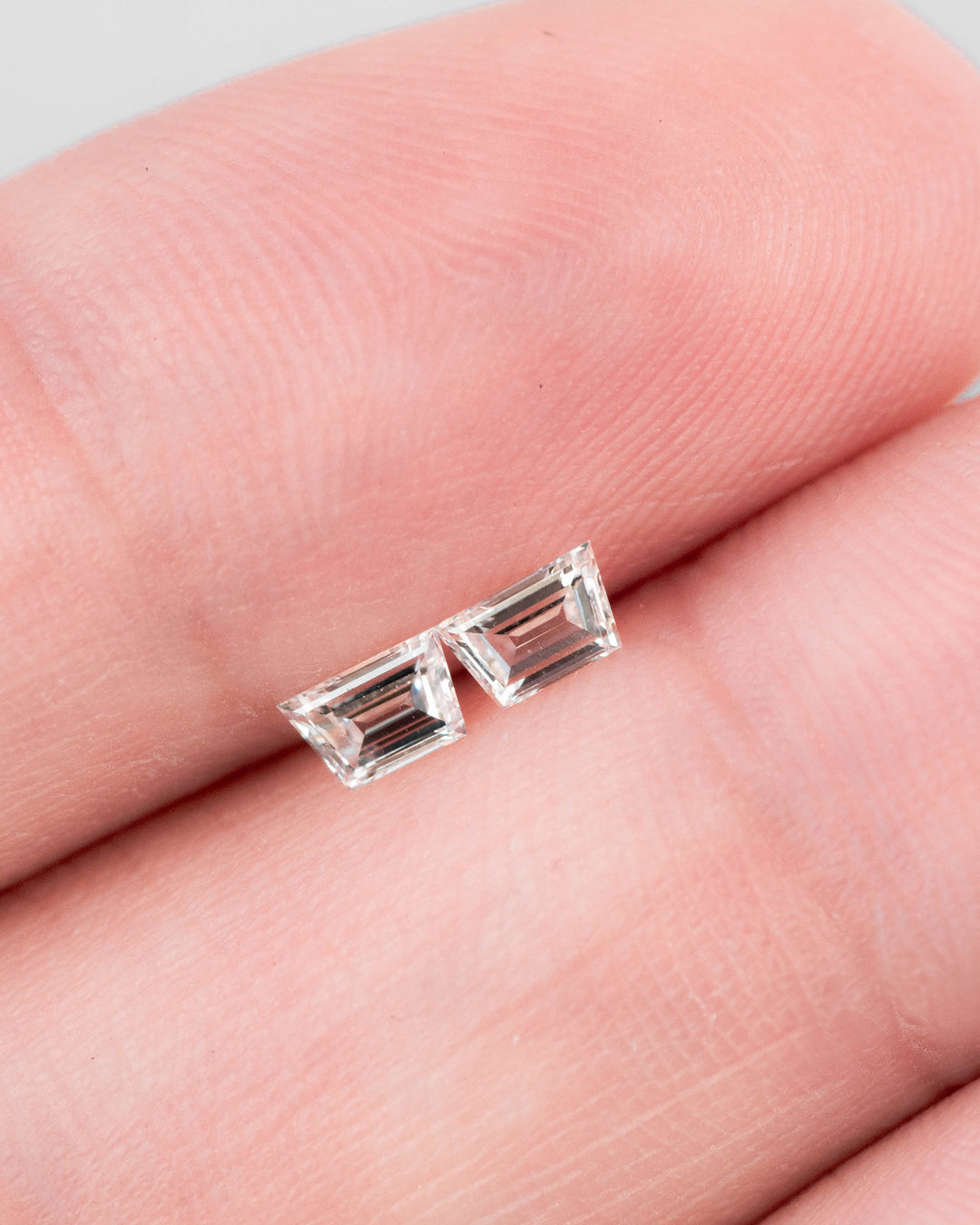 Matched pair of 0.37 carat G color VVS-VS trapezoid step cut diamonds resting on fingertips, showcasing the clean faceting, clarity, and precision symmetry of untreated South African diamonds.