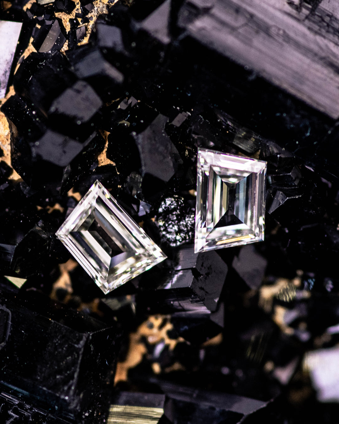 Macro photograph of 0.38 carat G–H color VVS clarity trapezoid step cut diamond pair from South Africa resting on black crystal matrix. The untreated diamonds showcase clean step faceting, sharp symmetry, and high brilliance.
