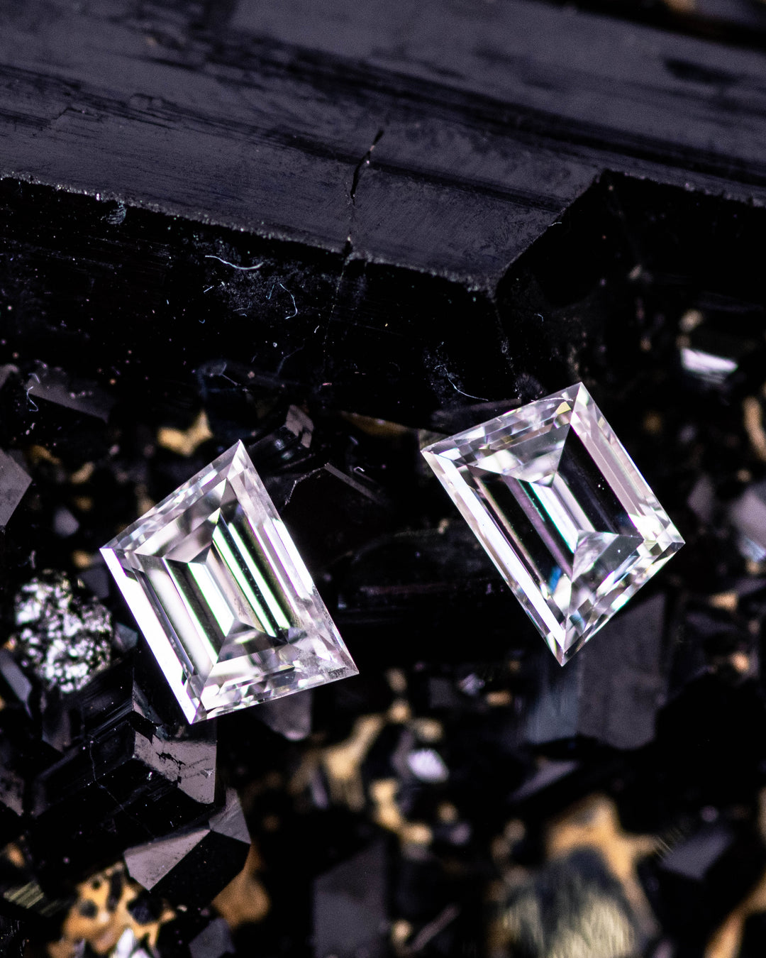 Macro photo of a 0.42 carat D color VVS clarity trapezoid step cut diamond pair from South Africa placed on a dark mineral matrix. The untreated diamonds exhibit crisp step facets, geometric precision, and mirror-like brilliance.