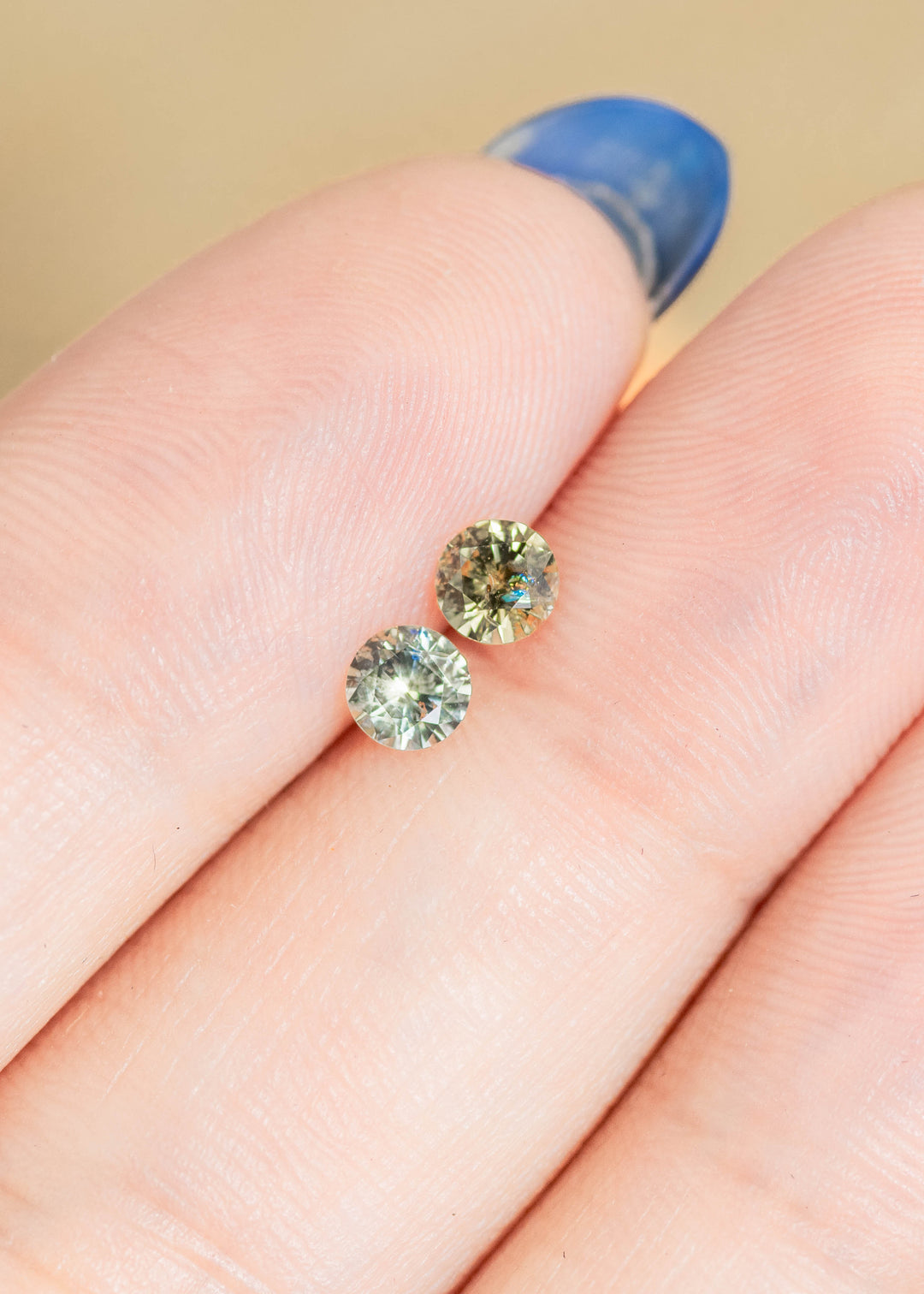 Loose matching pair of yellow, teal and green Montana sapphires resting on fingertips for scale, showing the round brilliant cut and natural color variation.