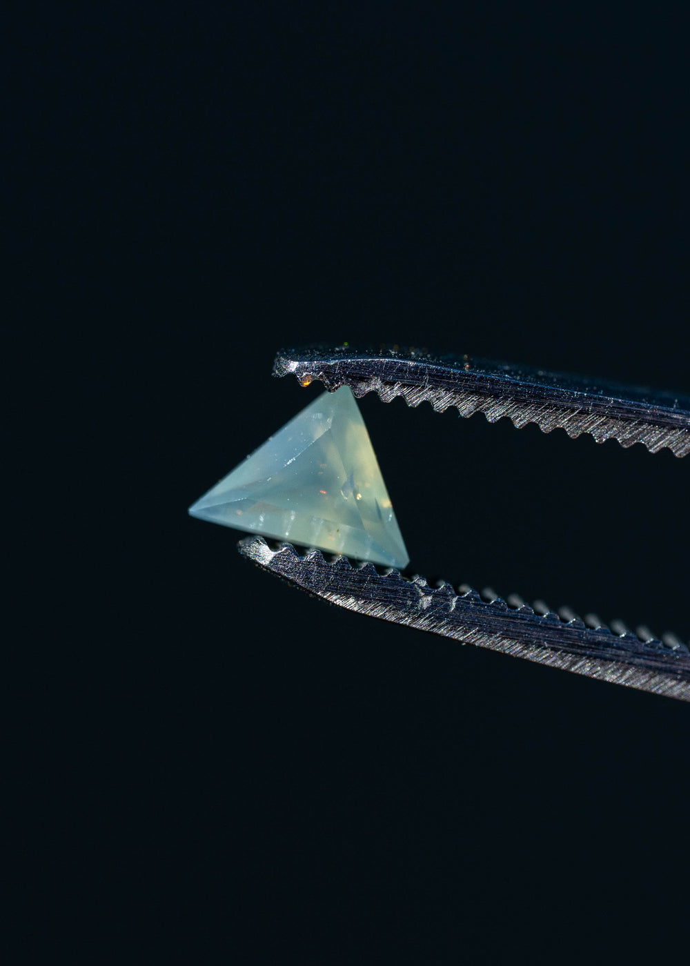 Close-up photograph of an untreated 0.62ct icy seafoam blue sapphire held in metal tweezers against a dark background, highlighting its triangular outline, translucent appearance and precision custom faceting.