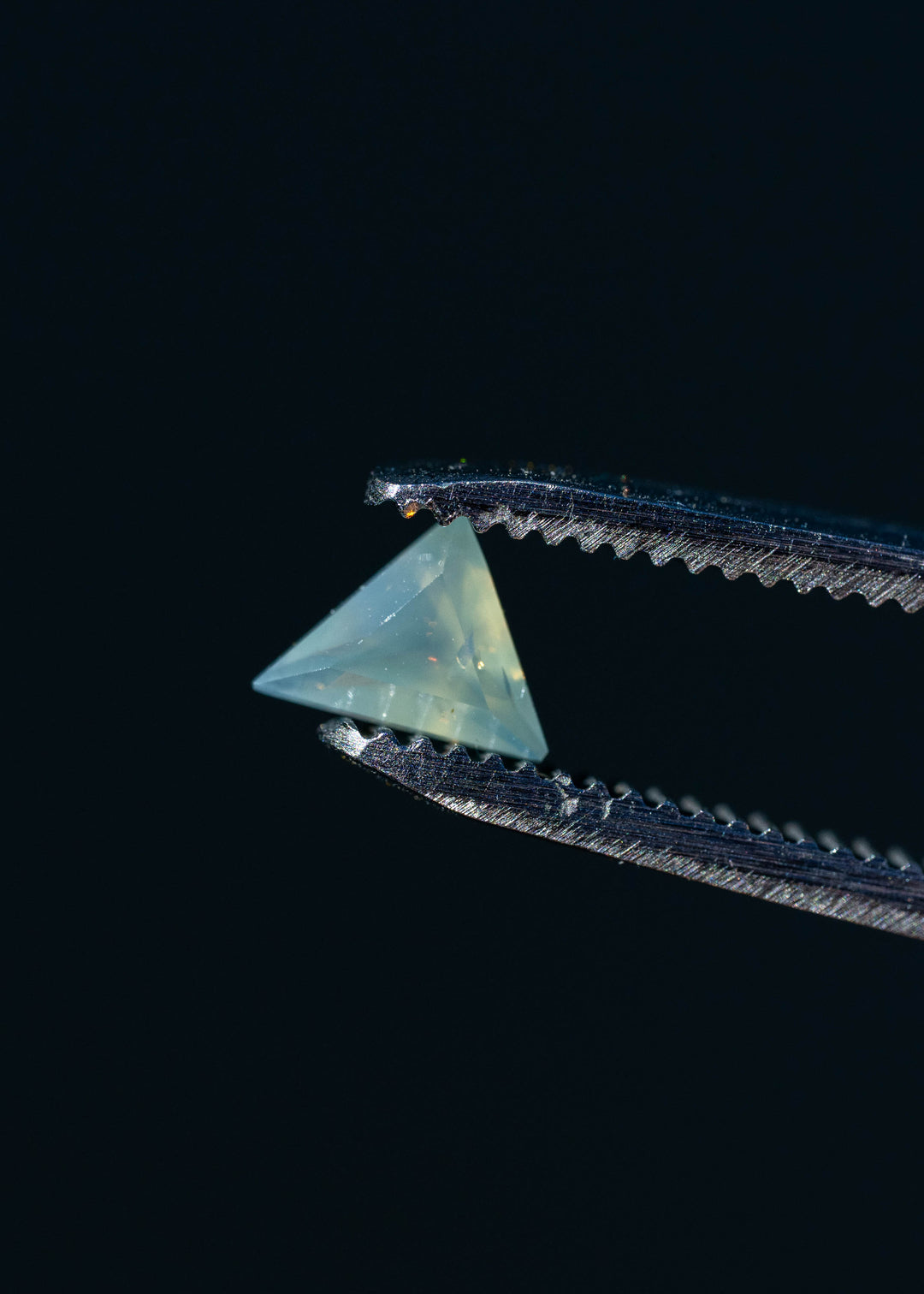 Close-up photograph of an untreated 0.62ct icy seafoam blue sapphire held in metal tweezers against a dark background, highlighting its triangular outline, translucent appearance and precision custom faceting.