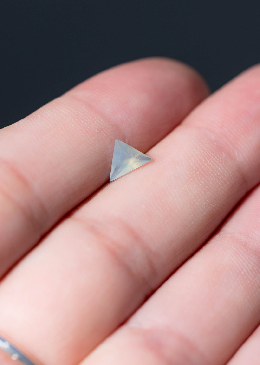 Untreated 0.62 carat opalescent icy seafoam blue sapphire from Nigeria photographed on fingers for scale. Its precision custom trillion cut measures 6.88 × 5.21 mm.