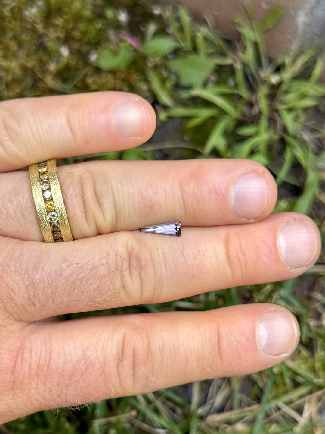Tanzanite gemstone resting on fingers, showcasing its custom elongated tapered baguette cut in multicolor hues of gray, peach, and violet.