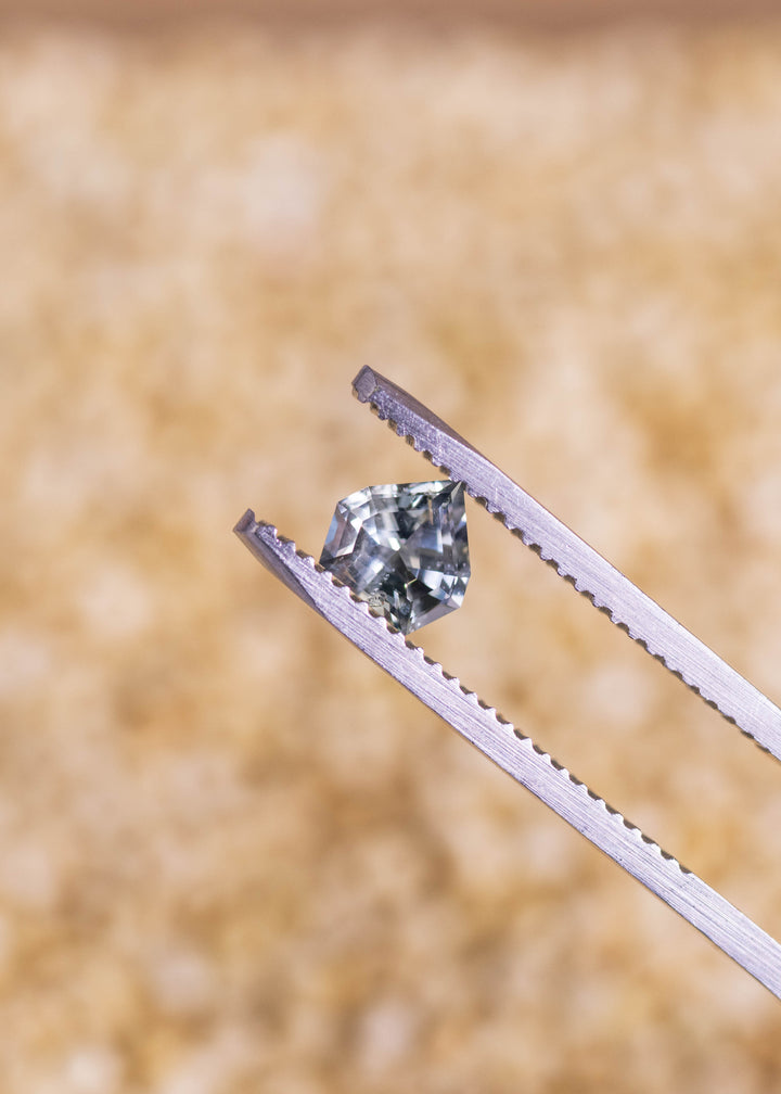 Loose green teal blue Montana sapphire held in tweezers, showing the geometric shield mix cut and clean faceting under natural light.