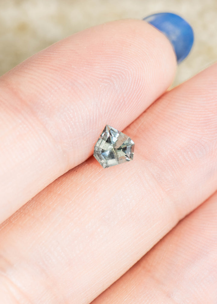 Loose green, teal and blue Montana sapphire resting on fingertip for scale, highlighting the geometric shield mix cut and natural color variation.