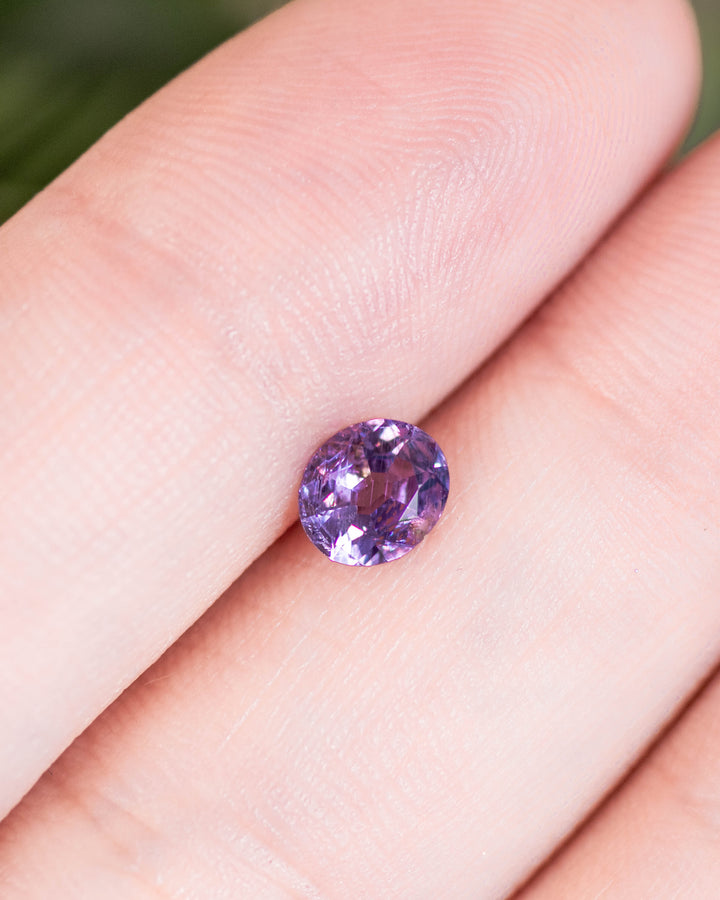 0.68 carat violet purple sapphire resting on fingertip, capturing its delicate brilliance and modified oval cut under natural light, untreated gem from Umba River Valley, Tanzania.