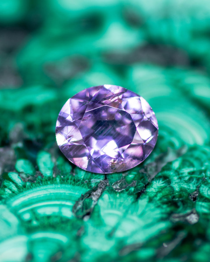 0.68 carat violet purple sapphire displayed on a rich green mineral surface, showing its modified oval cut brilliance and natural untreated color, ethically sourced from Umba River Valley, Tanzania.