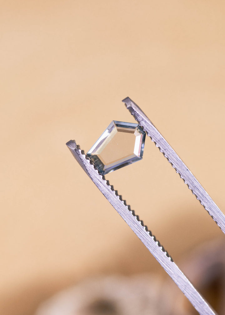 Loose green, blue and teal Montana sapphire held in tweezers, highlighting the thin portrait cut profile and clean pentagon shape under soft light.