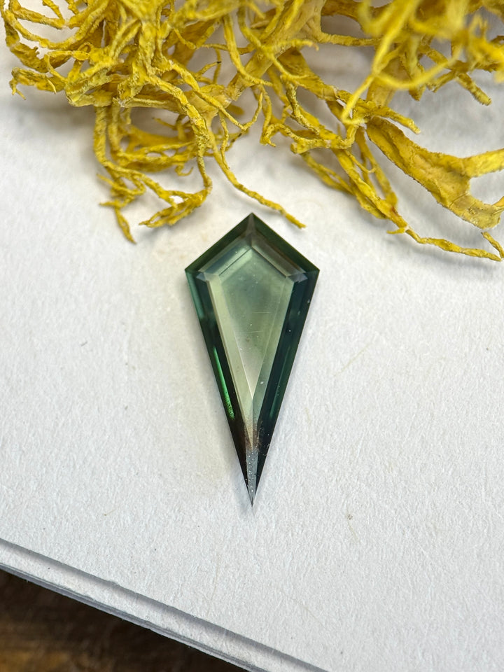 0.76 carat teal green sapphire kite portrait cut on neutral surface with organic elements, showcasing sharp geometric structure.