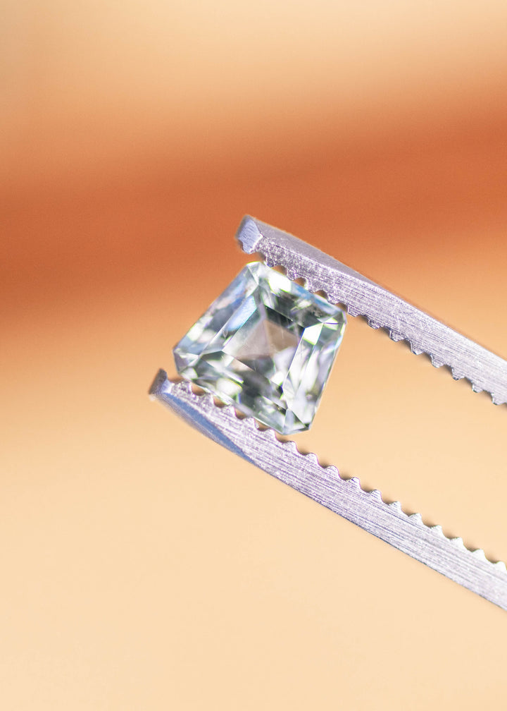 Seafoam step cut sapphire held in silver tweezers, revealing clean facet junctions, soft blue-green tone, and light reflections across the geometric crown.