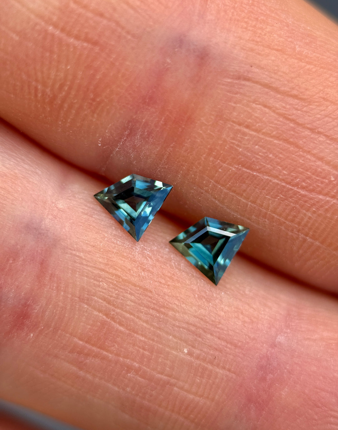 Pair of 0.84ct teal blue-green sapphires in kite step cut resting on fingertips, showing strong color zoning and clean precision faceting. Unheated sapphire pair from Queensland Gemfields, Australia.