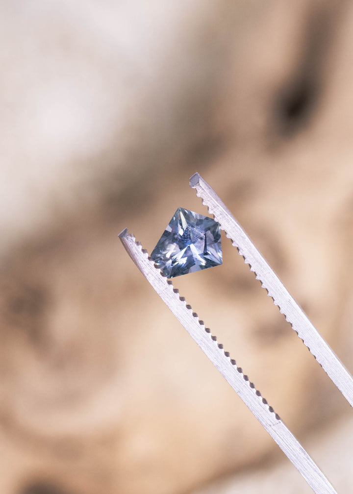 0.85ct blue green violet Montana sapphire held in jeweler’s tweezers, modified kite cut precision faceting reflecting light for custom jewelry design.
