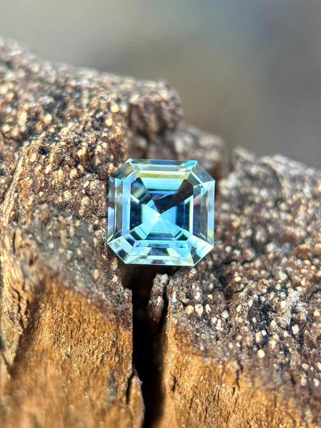 A square-cut light blue gemstone, likely a cushion-cut or emerald-cut sapphire, resting on a textured piece of weathered wood.