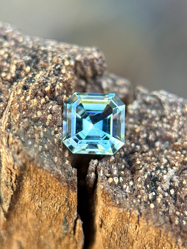 A square-cut light blue gemstone, likely a cushion-cut or emerald-cut sapphire, resting on a textured piece of weathered wood.