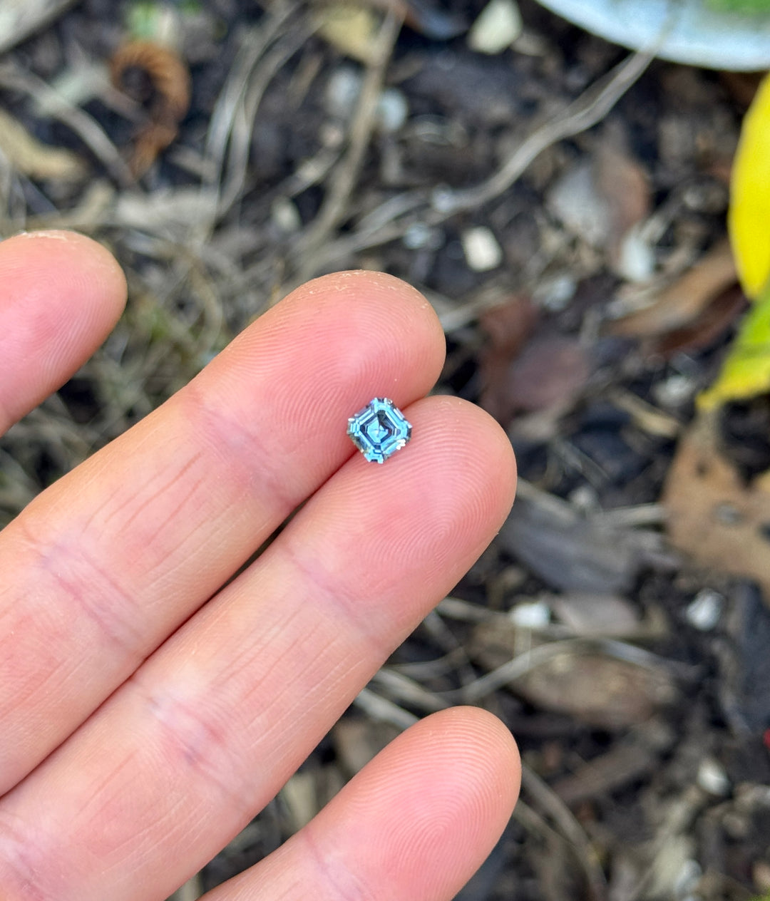 A tiny square-shaped light blue gemstone held between a person's fingers, with a blurry outdoor ground background of leaves and soil.