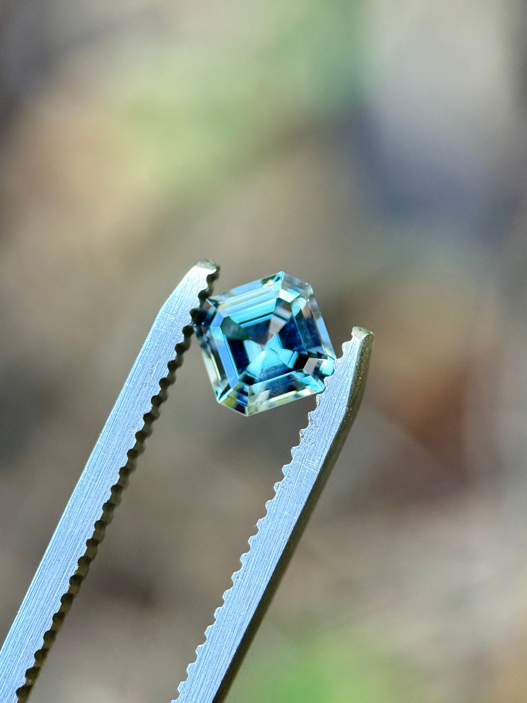 A loose square-cut light blue gemstone held between silver metal tweezers against a blurred natural background.
