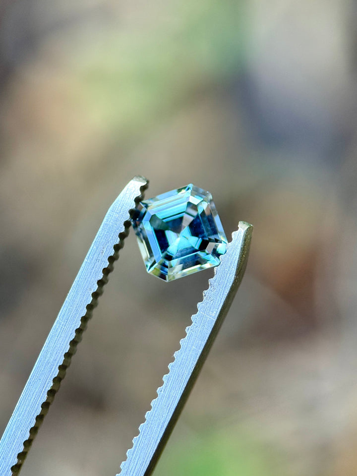 A loose square-cut light blue gemstone held between silver metal tweezers against a blurred natural background.