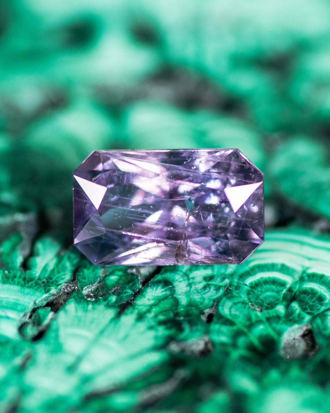 0.94 carat gray purple sapphire resting on a textured green mineral surface, displaying crisp facets and radiant reflections from its custom radiant cut, untreated sapphire from Umba River Valley, Tanzania.