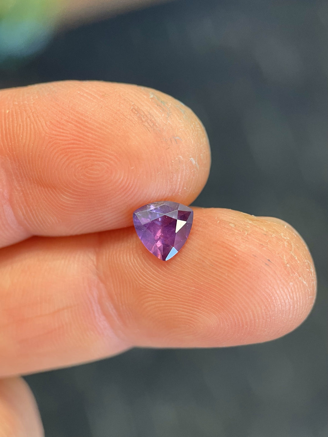 Close-up of a 0.94-carat custom step cut trillion sapphire in pink and purple hues, held gently on a fingertip with a natural background.