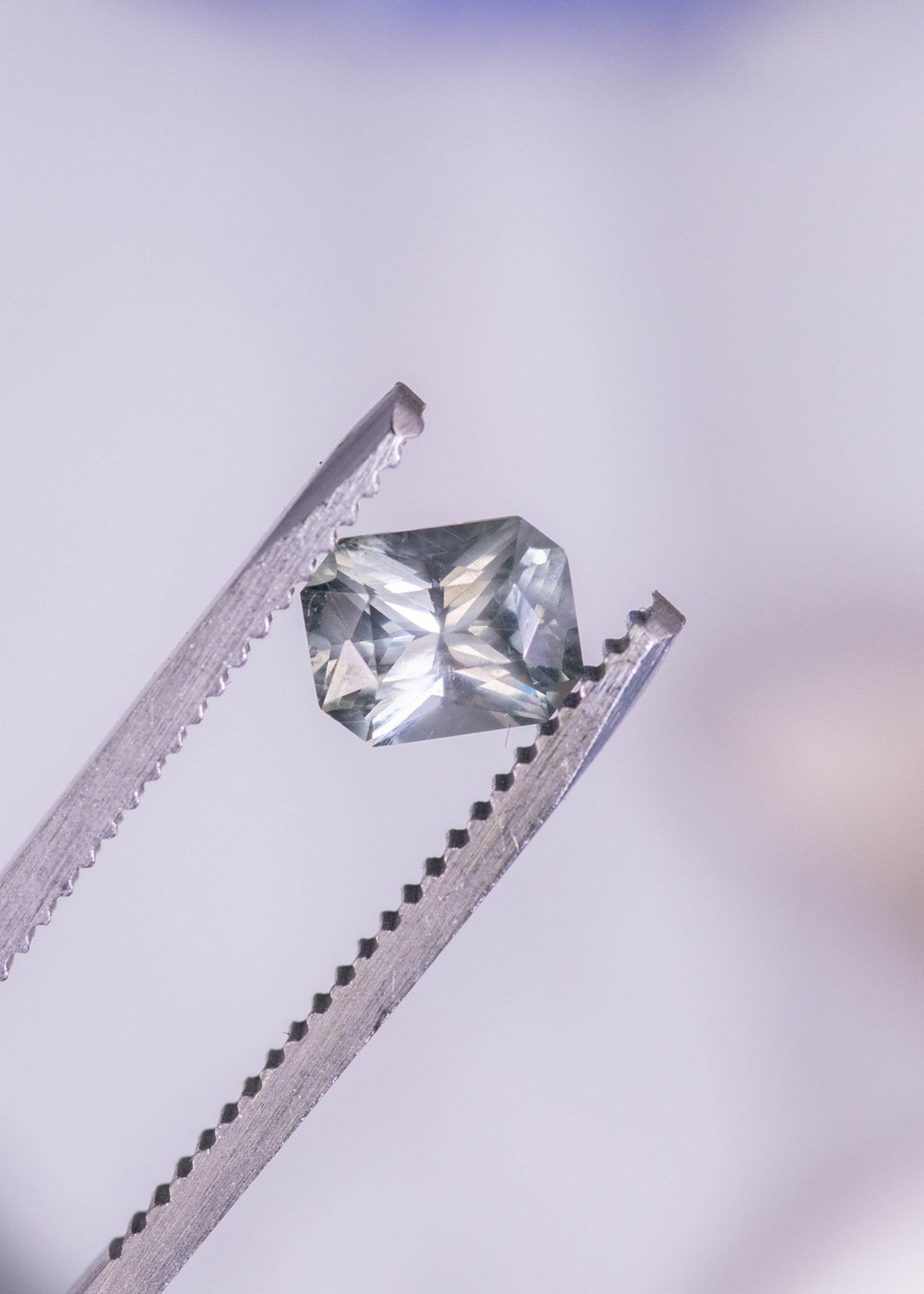 0.96 carat teal gray blue Montana sapphire mixed radiant cut held in gem tweezers, displaying step and brilliant facets with crisp geometric symmetry and natural color shift.