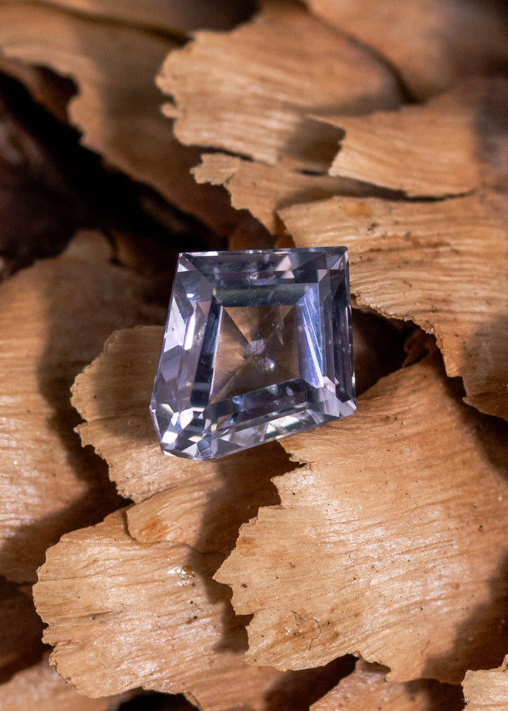 0.98 carat unheated Montana sapphire resting on natural wood, gray violet teal custom modified kite step cut from El Dorado Bar Deposit showing geometric step faceting
