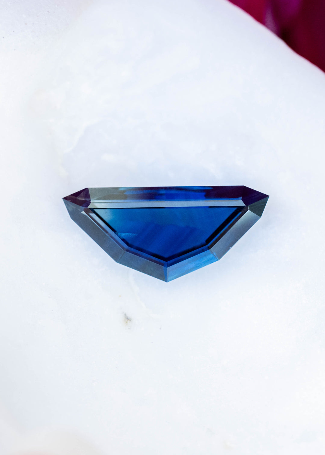 Elongated, angular kite-shaped sapphire resting horizontally on a white flower petal. Its broad center is saturated blue, with darker navy areas and pale blue-gray step-cut facets around the edges.