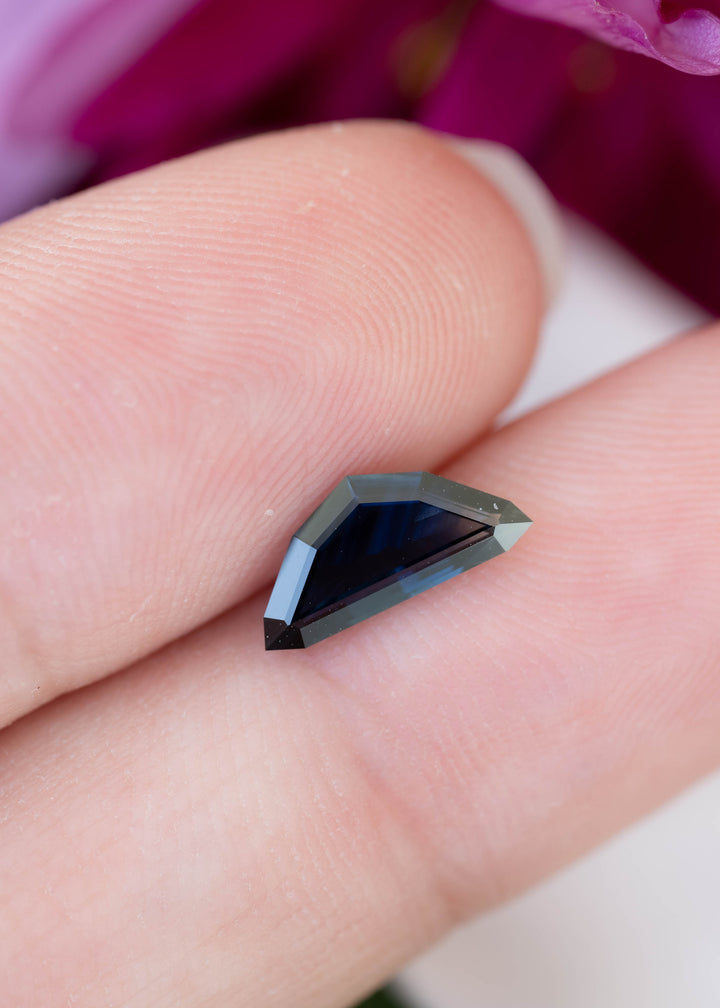 Small, elongated kite-shaped sapphire resting horizontally across two fingertips. Its center appears dark navy blue, bordered by muted blue-gray facets and a sharply pointed end.