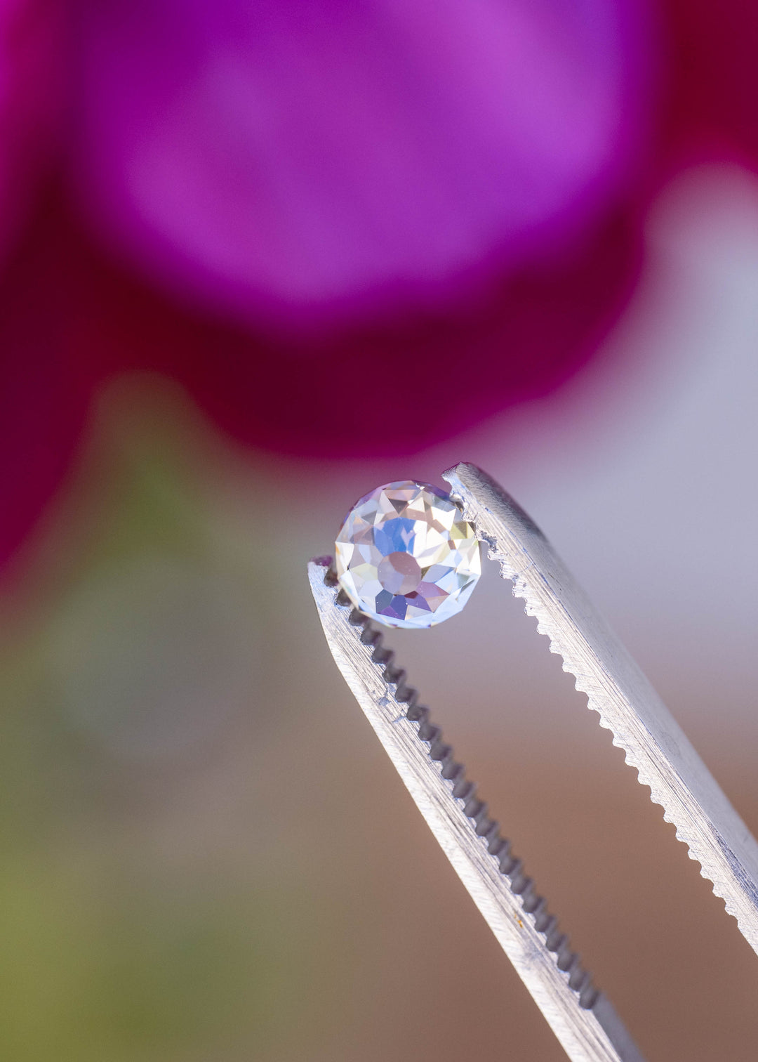 0.99 carat Mughal cut diamond held in jeweler’s tweezers against a softly blurred pink floral background, showing its round outline and antique facet pattern.