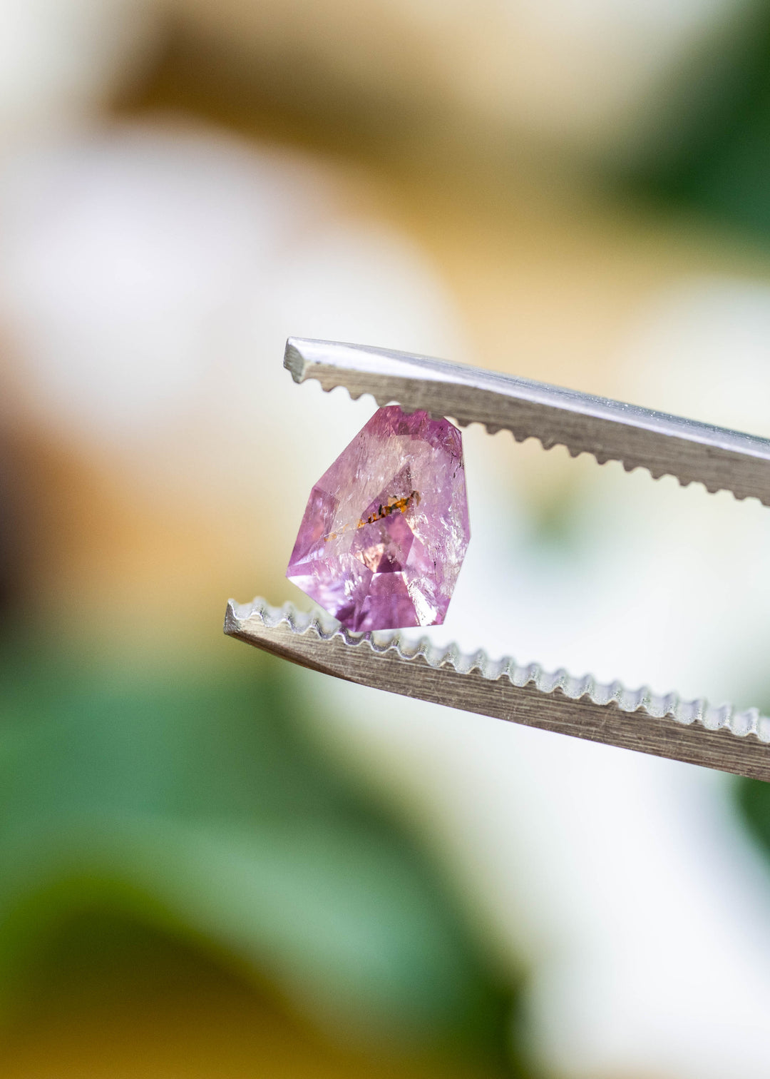 Close-up of a natural untreated 1.01 carat Colombian sapphire in tweezers, highlighting its custom shield mix cut, soft rose pink color, and natural mineral inclusion.