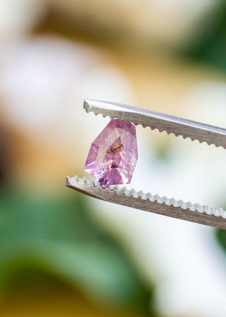 Close-up of a natural untreated 1.01 carat Colombian sapphire in tweezers, highlighting its custom shield mix cut, soft rose pink color, and natural mineral inclusion.