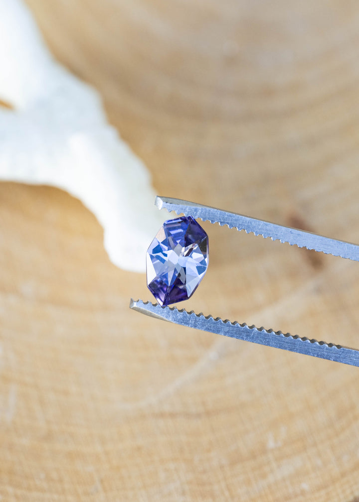 Close-up of a natural 1.11 carat Merelani Hills tanzanite in tweezers showcasing its precision custom geometric marquise cut and pleochroic purple, teal, and gray color.