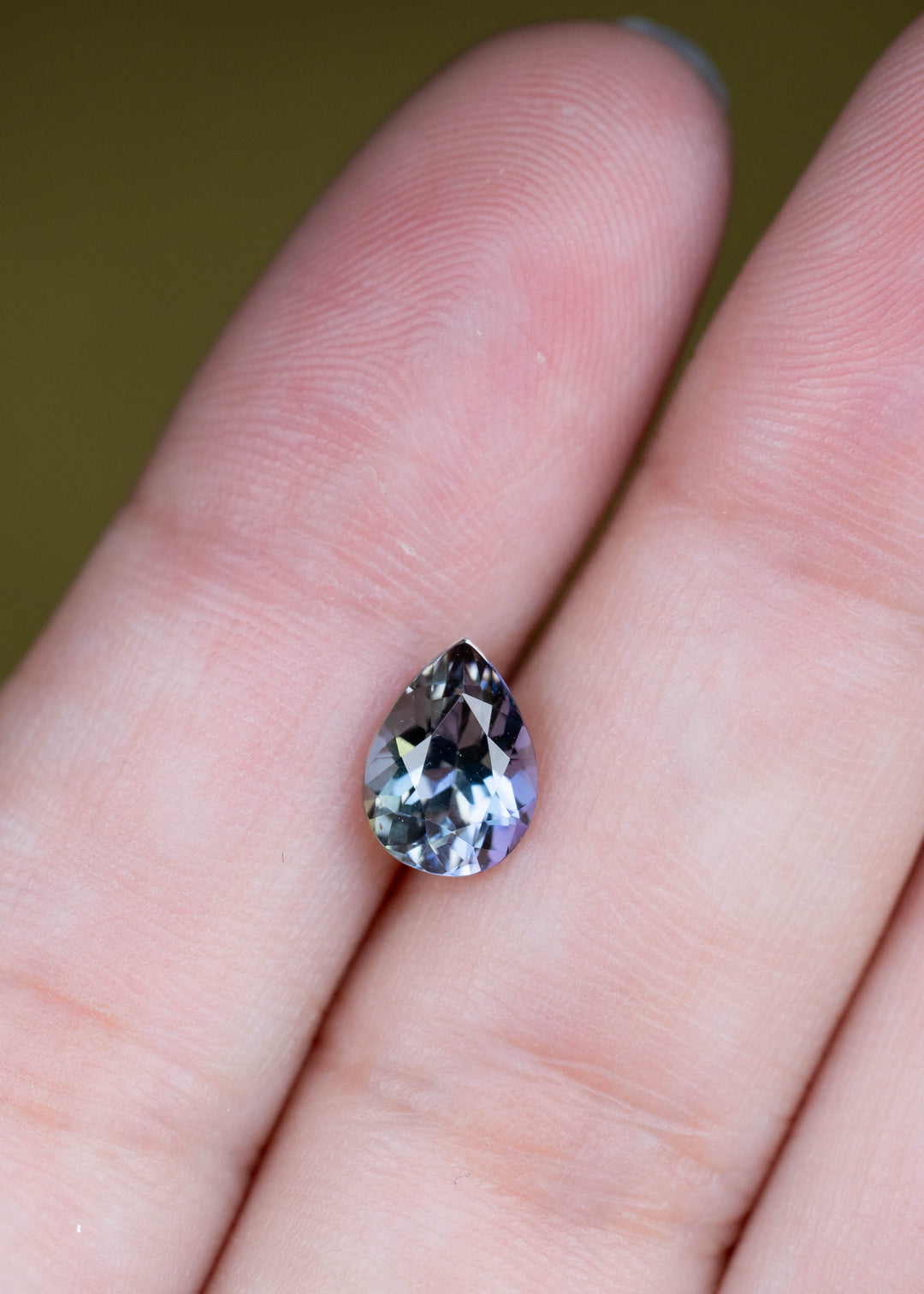 Natural 1.55 carat Merelani tanzanite shown on fingertips, displaying a precision modified mixed brilliant pear cut with complex mermaid coloration.