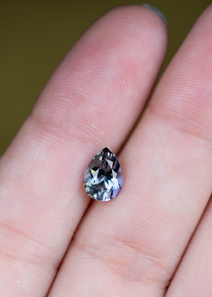 Natural 1.55 carat Merelani tanzanite shown on fingertips, displaying a precision modified mixed brilliant pear cut with complex mermaid coloration.