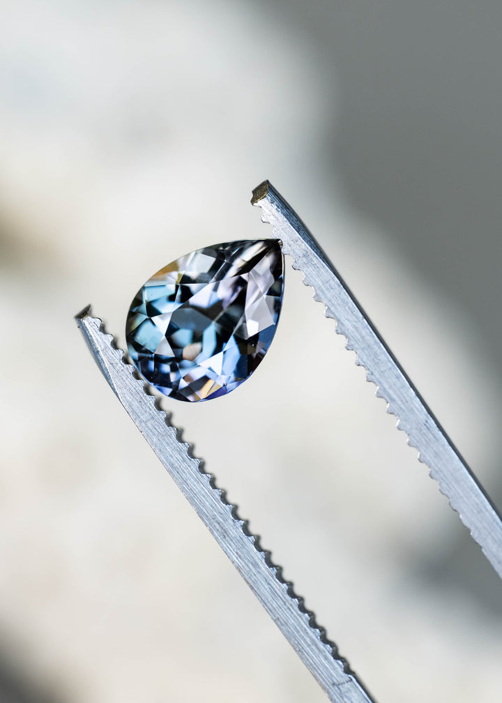 Close-up of a natural 1.55 carat untreated pear cut tanzanite in tweezers highlighting exceptional pleochroic blue, teal, green, purple, and yellow hues.