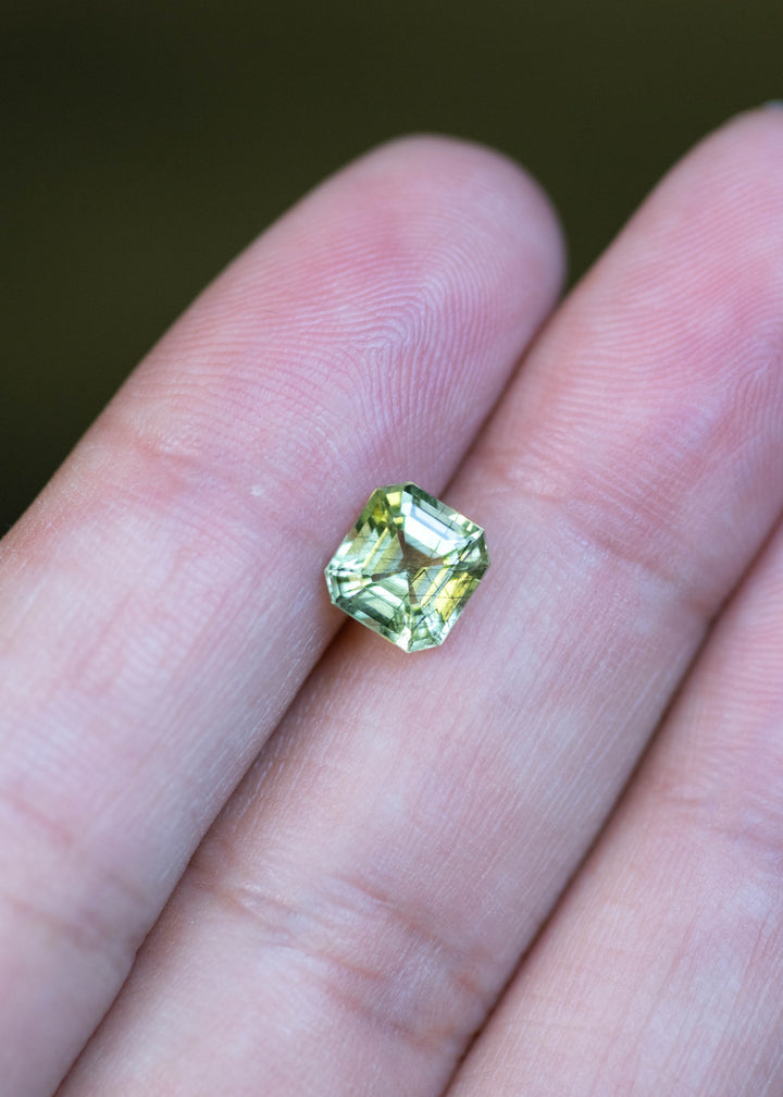 Natural lime green peridot from Pakistan displayed on fingertips for scale, showcasing its custom Asscher cut, untreated crystal, and vibrant green brilliance.